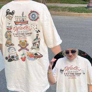 May include: Cream-colored t-shirt featuring a tattoo-style design with the text "Walt's Ink & Paint Tattoo Shop." The back of the shirt displays various tattoo-inspired illustrations, including a skull, boat, and snake.