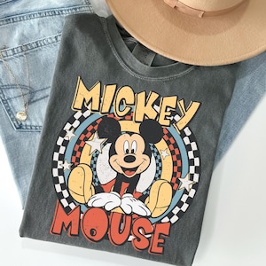 May include: A white ceramic mug with a black handle and the text "But First, Coffee" in a bold, black font. The mug is set against a blurred background. The mug appears to be approximately 10 cm tall.