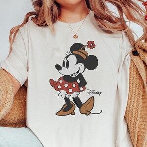 Może przedstawiać: Kremowy t-shirt z grafiką Myszki Minnie. Myszka Minnie jest przedstawiona w swojej kultowej czerwonej sukience w kropki, brązowych butach i kwiatkiem na kapeluszu. Poniżej znajduje się logo Disney.