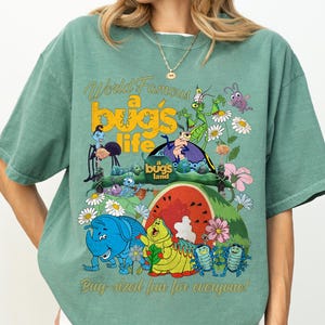 May include: Green t-shirt featuring a colorful graphic from "a bug's life". The design includes cartoon insects, flowers, and a watermelon slice. Text on the shirt reads "World Famous a bug's life" and "Bug-sized fun for everyone!"