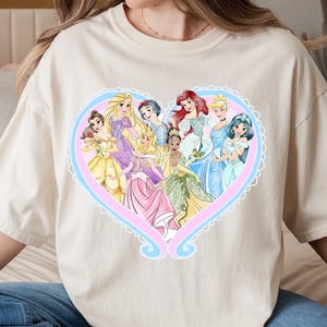 May include: Cream-colored t-shirt featuring a heart-shaped design with illustrations of various princesses in colorful gowns. The heart is outlined in pink and blue with a decorative border. The shirt is made of a soft material.