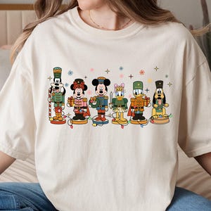 May include: Cream-colored t-shirt featuring a festive design with cartoon characters dressed as nutcrackers. The characters include Mickey Mouse, Minnie Mouse, Donald Duck, and Goofy, with colorful details and holiday lights. A fun and whimsical holiday tee.