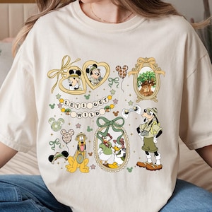 May include: Cream-colored t-shirt featuring various Disney characters and the phrase "LET'S GET WILD." The design includes Mickey and Minnie Mouse, Goofy, Donald and Daisy Duck, and Pluto, with safari-themed elements and heart-shaped accents.