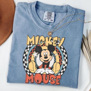 May include: A blue Comfort Colors t-shirt with a Mickey Mouse graphic. The design features the words "Mickey Mouse" in yellow and red, with a checkered and star-filled background. A gold necklace with a pendant is placed on the shirt.
