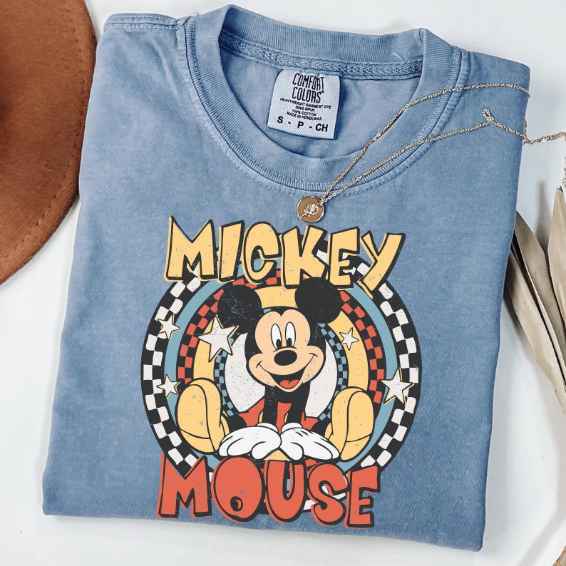 May include: A blue Comfort Colors t-shirt with a Mickey Mouse graphic. The design features the words "Mickey Mouse" in yellow and red, with a checkered and star-filled background. A gold necklace with a pendant is placed on the shirt.