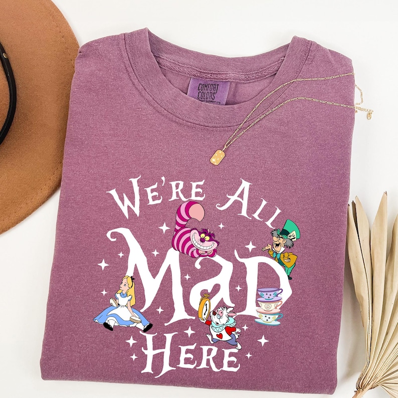 May include: A mauve-colored t-shirt featuring the phrase "We're All Mad Here" in white script, with illustrations of characters from Alice in Wonderland. A gold necklace with a rectangular pendant is draped across the shirt.