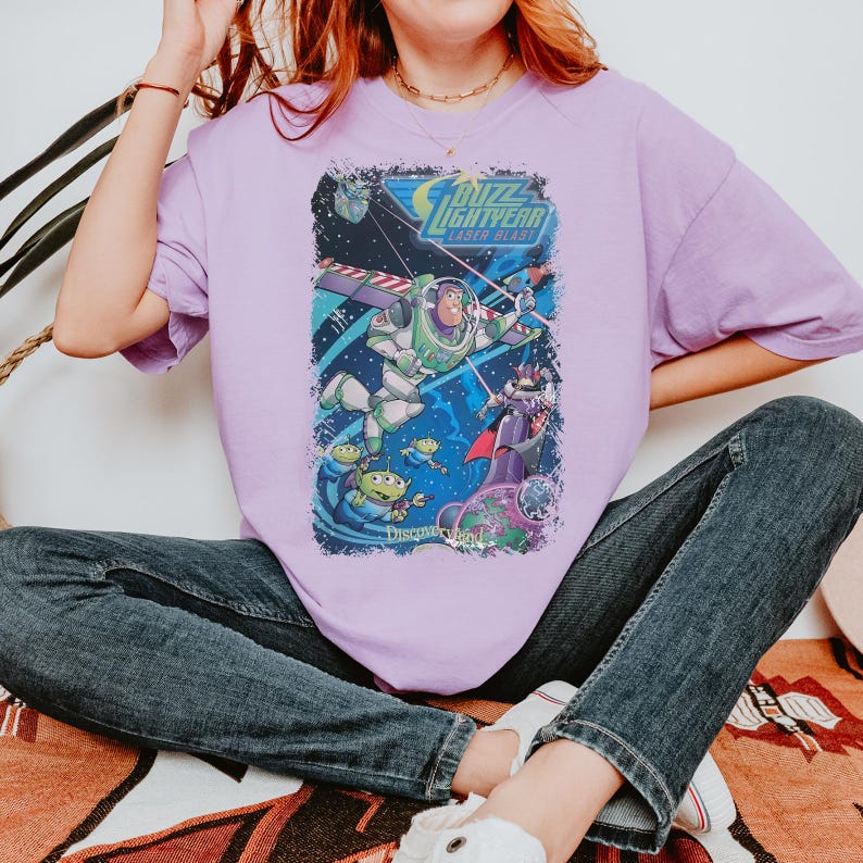 K&ouml;nnte beinhalten: Ein helllila T-Shirt mit einer Grafik aus dem Film "Buzz Lightyear Laser Blast" mit Buzz Lightyear, Aliens und Zurg. Das Disneyland-Logo befindet sich unten. Das T-Shirt besteht aus einem weichen Material.