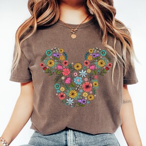 K&ouml;nnte beinhalten: Ein braunes T-Shirt mit einem floralen Mickey-Mouse-Design. Das Design besteht aus bunten Blumen in Rosa-, Gelb-, Blau- und Lilat&ouml;nen. Das Shirt hat kurze &Auml;rmel und einen Rundhalsausschnitt.