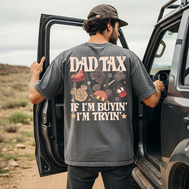 May include: A gray t-shirt with the text "DAD TAX" and "IF I'M BUYIN' I'M TRYIN'" in a vintage style. The back of the shirt features cartoon images of Mickey Mouse and treats. The person is standing next to a vehicle.