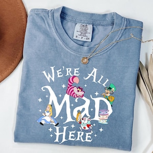 May include: A blue Comfort Colors t-shirt featuring the phrase "We're All Mad Here" with illustrations of characters from Alice in Wonderland. The shirt has a crew neck and short sleeves. A gold necklace with a pendant is draped across the shirt.