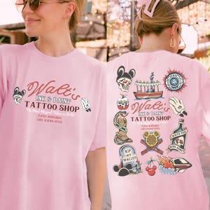 May include: Pink t-shirt with a tattoo shop design. The front features the text "Walt's Ink & Paint Tattoo Shop" with Mickey Mouse-inspired graphics. The back has various tattoo flash designs, including a skull, snake, and ship.