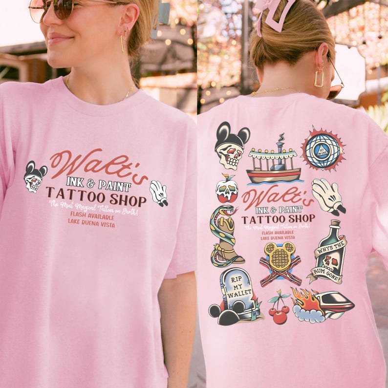 May include: Pink t-shirt with a tattoo shop design. The front features the text "Walt's Ink & Paint Tattoo Shop" with Mickey Mouse-inspired graphics. The back has various tattoo flash designs, including a skull, snake, and ship.