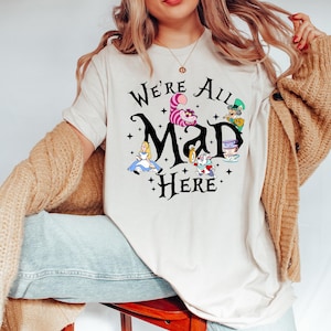 May include: A cream-colored t-shirt featuring the phrase "We're All Mad Here" in black script, with illustrations of characters from Alice in Wonderland. The design includes Alice, the Cheshire Cat, the Mad Hatter, and the White Rabbit.