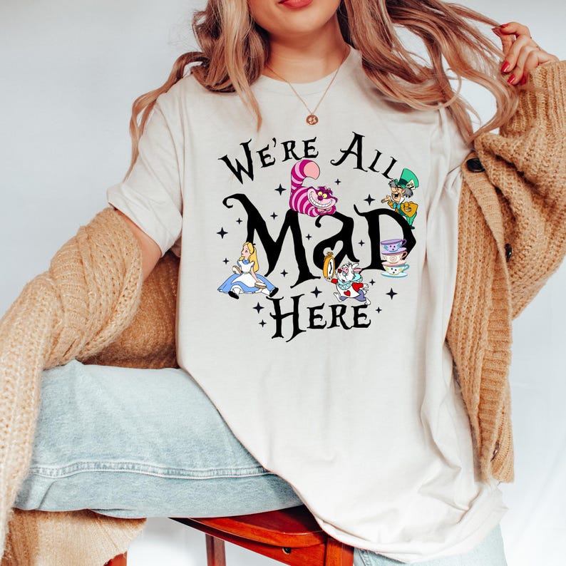 May include: A cream-colored t-shirt featuring the phrase "We're All Mad Here" in black script, with illustrations of characters from Alice in Wonderland. The design includes Alice, the Cheshire Cat, the Mad Hatter, and the White Rabbit.