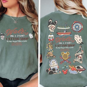 May include: A sage green t-shirt with the text "Walt's Ink & Paint Tattoo Shop" in red and black. The shirt features various tattoo-style graphics, including a skull, apple, snake, and a tombstone that says "RIP MY WALLET."