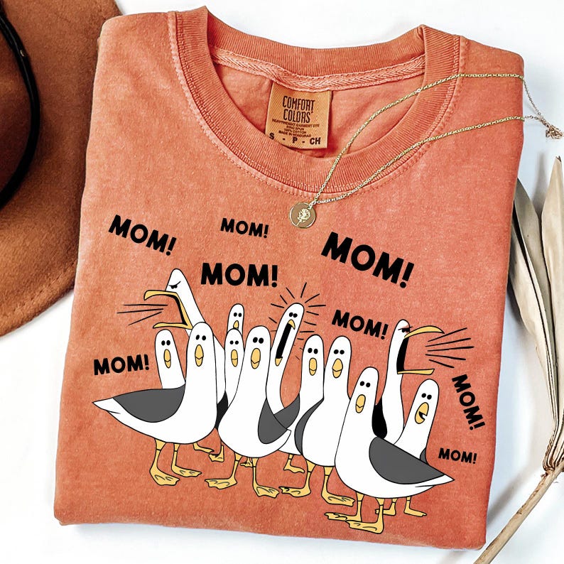 May include: A coral-colored t-shirt with the word "MOM!" repeated around a cartoon illustration of seagulls. The seagulls are white with gray wings and yellow feet. A gold necklace with a pendant is on the shirt.