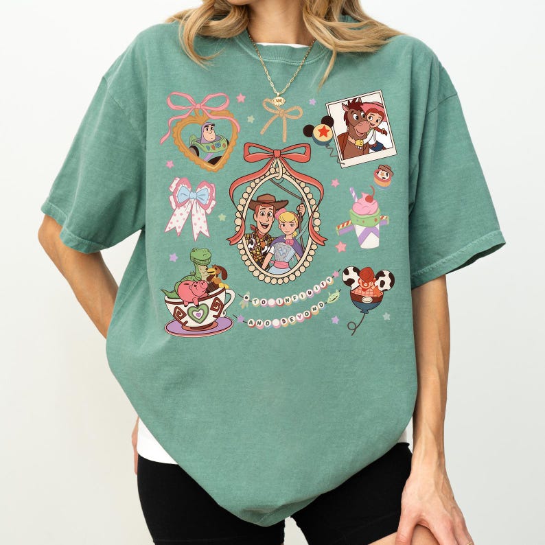 May include: Sage green t-shirt with cartoon graphics of characters from a popular animated film. The design includes a heart-shaped Buzz Lightyear, a photo of Woody and Jessie, and other playful illustrations.