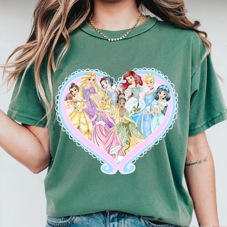 May include: Green t-shirt featuring a heart-shaped design with illustrations of various princesses. The heart is outlined in white with a pink and blue interior. The princesses are depicted in their signature gowns.