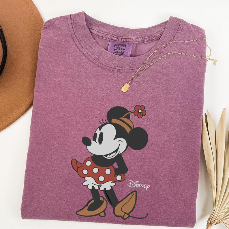 Może przedstawiać: Fioletowa koszulka Comfort Colors z grafiką Myszki Minnie. Postać z kresk&oacute;wki ma na sobie czerwoną i białą sukienkę w kropki, brązowe buty i pasujący kapelusz. Złoty naszyjnik z małą zawieszką spoczywa na koszulce.