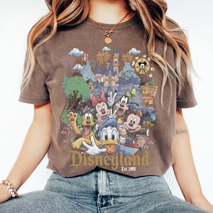 May include: A brown short-sleeve t-shirt featuring a graphic print of Disney characters including Mickey Mouse, Minnie Mouse, Donald Duck, Goofy, and Pluto in front of a castle. The text Disneyland Est. 1955 is printed in gold at the bottom.