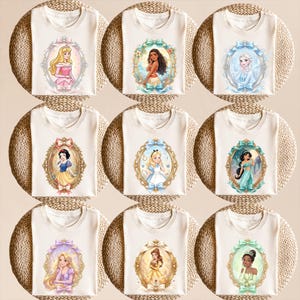 Op de afbeelding: Negen cr&egrave;mekleurige t-shirts, elk met een prinsessenportret in een sierlijke, ovale lijst. De prinsessen zijn Aurora, Moana, Elsa, Sneeuwwitje, Alice, Jasmine, Rapunzel, Belle en Tiana. De lijsten hebben gouden en pastelkleurige accenten.