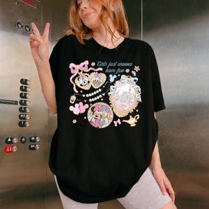 May include: Black t-shirt featuring a colorful graphic design with the text "Girls just wanna have fun." The design includes cartoon characters, bows, and a castle. The shirt has a classic crew neck and short sleeves.