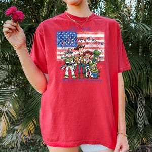 May include: Red t-shirt featuring a graphic of the American flag and cartoon characters. The shirt has the text "1776-2026" and "America's 250th Anniversary" in blue. The design includes fireworks and patriotic elements.