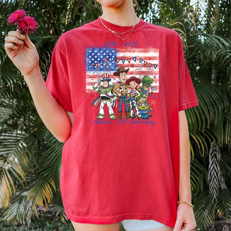 May include: Red t-shirt featuring a graphic of the American flag and cartoon characters. The shirt has the text "1776-2026" and "America's 250th Anniversary" in blue. The design includes fireworks and patriotic elements.