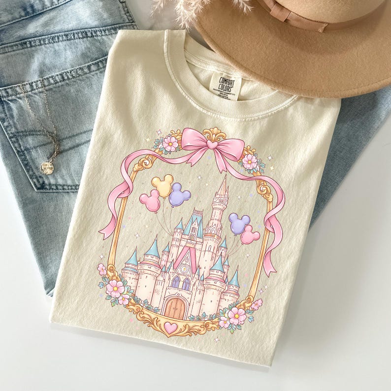 May include: Cream-colored t-shirt featuring a fairytale castle design within a pink ribbon frame, adorned with Mickey Mouse balloons and floral accents. The shirt has the text "COMFORT COLORS" on the tag. The shirt is folded on top of blue jeans.