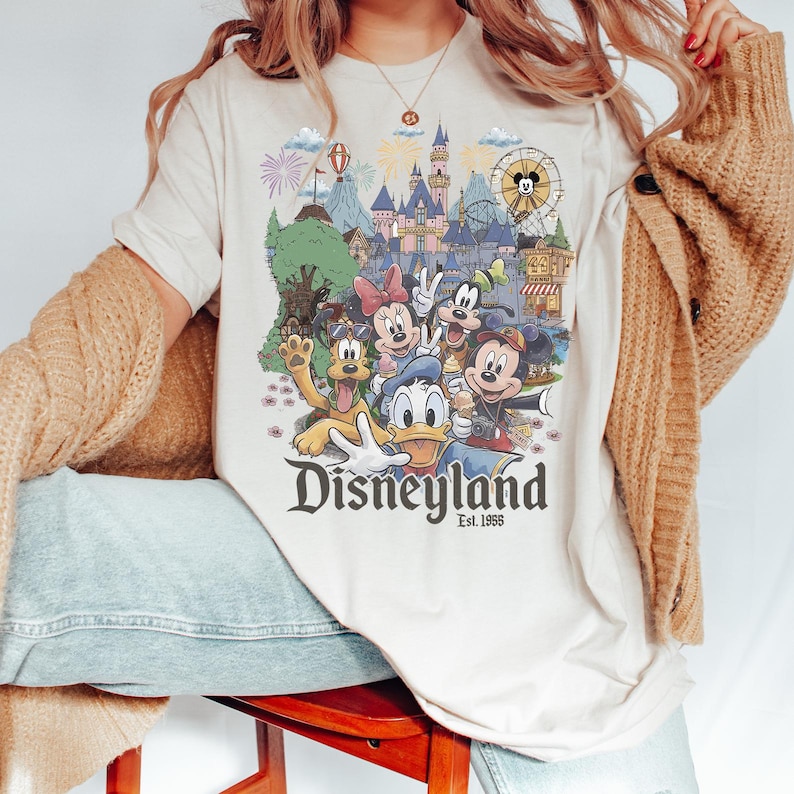 May include: A cream-colored t-shirt featuring a colorful graphic of Disney characters including Mickey Mouse, Minnie Mouse, Donald Duck, Goofy, and Pluto in front of a castle, with the text Disneyland Est. 1955 printed below the characters.