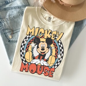 May include: Cream-coloured t-shirt with a Mickey Mouse graphic. The design features the words "Mickey Mouse" in yellow and red, set against a circular background of stars and a chequered pattern. The shirt is folded with blue jeans and a tan hat.