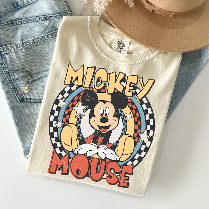 May include: Cream-coloured t-shirt with a Mickey Mouse graphic. The design features the words "Mickey Mouse" in yellow and red, set against a circular background of stars and a chequered pattern. The shirt is folded with blue jeans and a tan hat.
