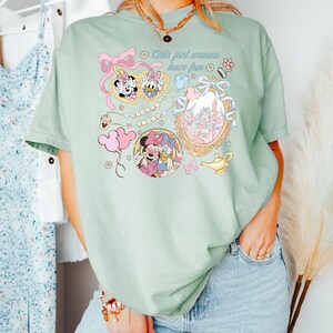 May include: A light green t-shirt featuring a colorful graphic design with the text "Girls just wanna have fun." The design includes illustrations of Minnie Mouse, Daisy Duck, and a castle, along with other decorative elements.