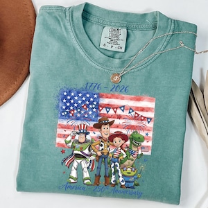 May include: A teal Comfort Colors t-shirt featuring a graphic of the American flag with fireworks and cartoon characters. The text "1776-2026" and "America 250th Anniversary" are printed on the shirt. A gold necklace is draped across the shirt.