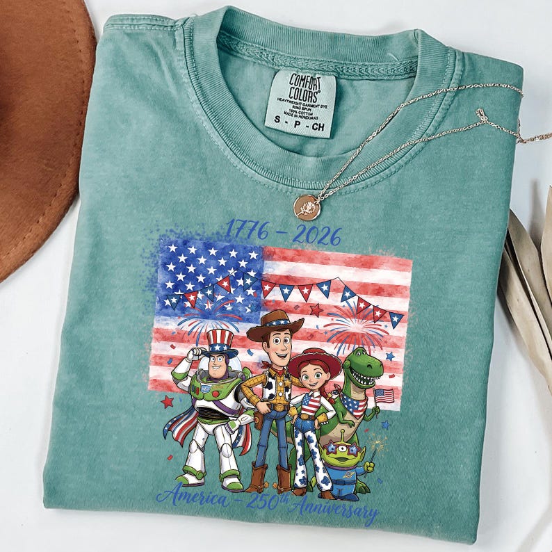 May include: A teal Comfort Colors t-shirt featuring a graphic of the American flag with fireworks and cartoon characters. The text "1776-2026" and "America 250th Anniversary" are printed on the shirt. A gold necklace is draped across the shirt.
