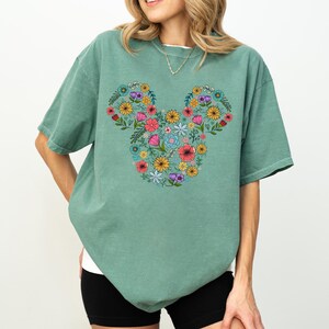 K&ouml;nnte beinhalten: Salbeigr&uuml;nes T-Shirt mit einem floralen Mickey-Mouse-Design. Das Design besteht aus verschiedenen bunten Blumen, darunter Rosa, Gelb und Blau, die in Form des Mickey-Mouse-Kopfes angeordnet sind. Das T-Shirt hat kurze &Auml;rmel und eine lockere Passform.