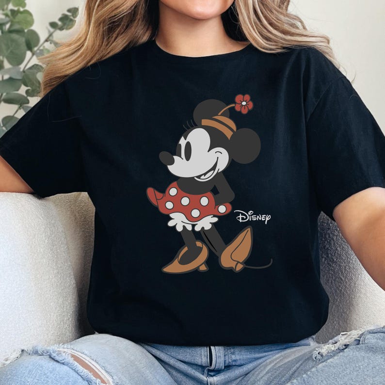 Może przedstawiać: Czarny t-shirt z kresk&oacute;wkowym wzorem Myszki Minnie. Myszka Minnie ma na sobie czerwoną sukienkę w kropki, brązowe buty i kwiat na kapeluszu. Logo Disneya jest nadrukowane na koszulce.