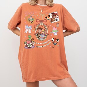 May include: Orange t-shirt with cartoon graphics of animated film characters. The design includes framed images, bows, and other decorative elements. The shirt is a comfortable, casual style.