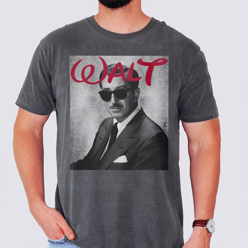 May include: Dark gray t-shirt featuring a black and white image of a man wearing sunglasses and a suit. The word "WALT" is written in red cursive above the image. The shirt has a crew neck and short sleeves.