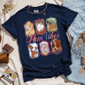 May include: Navy blue t-shirt featuring framed images of animated mothers and their babies. The shirt has the words "Mom Vibes" in a colorful script across the front. A gold necklace is draped on the shirt.