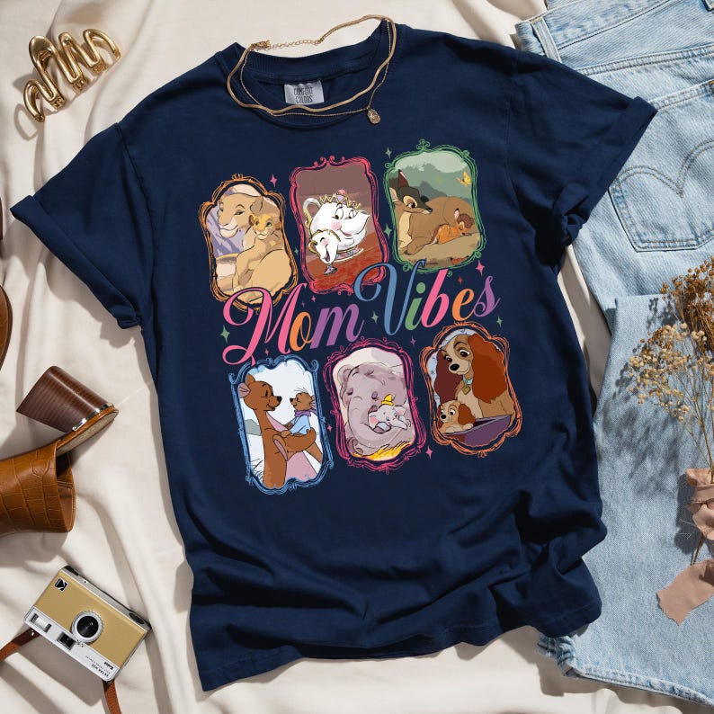 May include: Navy blue t-shirt featuring framed images of animated mothers and their babies. The shirt has the words "Mom Vibes" in a colorful script across the front. A gold necklace is draped on the shirt.