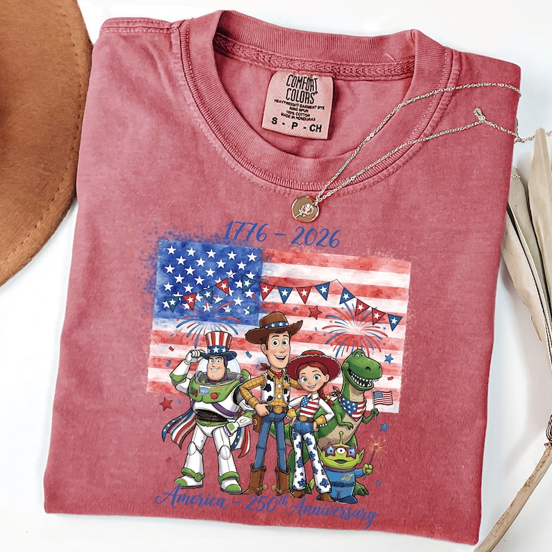 May include: A red t-shirt featuring a graphic of the American flag and characters from the movie Toy Story. The shirt has the text "1776-2026" and "America 250th Anniversary". A silver necklace with a pendant is on the shirt.