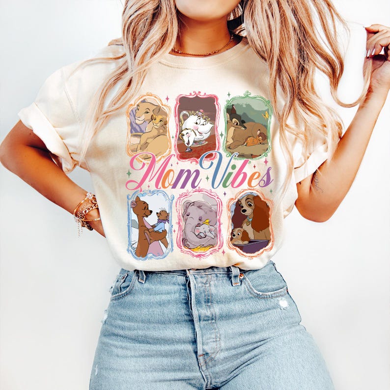 May include: Cream-colored t-shirt featuring the text "Mom Vibes" in a colorful script. The shirt displays framed illustrations of animated mothers and their children. The design includes images of lions, elephants, and dogs.
