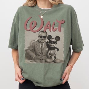 May include: Olive green t-shirt featuring a vintage-style graphic of Walt Disney with Mickey Mouse. The graphic includes the word "Walt" in a cursive font. The shirt has a relaxed fit and short sleeves.