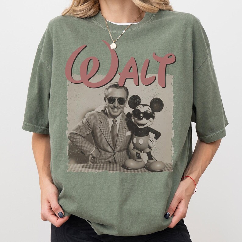 May include: Olive green t-shirt featuring a vintage-style graphic of Walt Disney with Mickey Mouse. The graphic includes the word "Walt" in a cursive font. The shirt has a relaxed fit and short sleeves.
