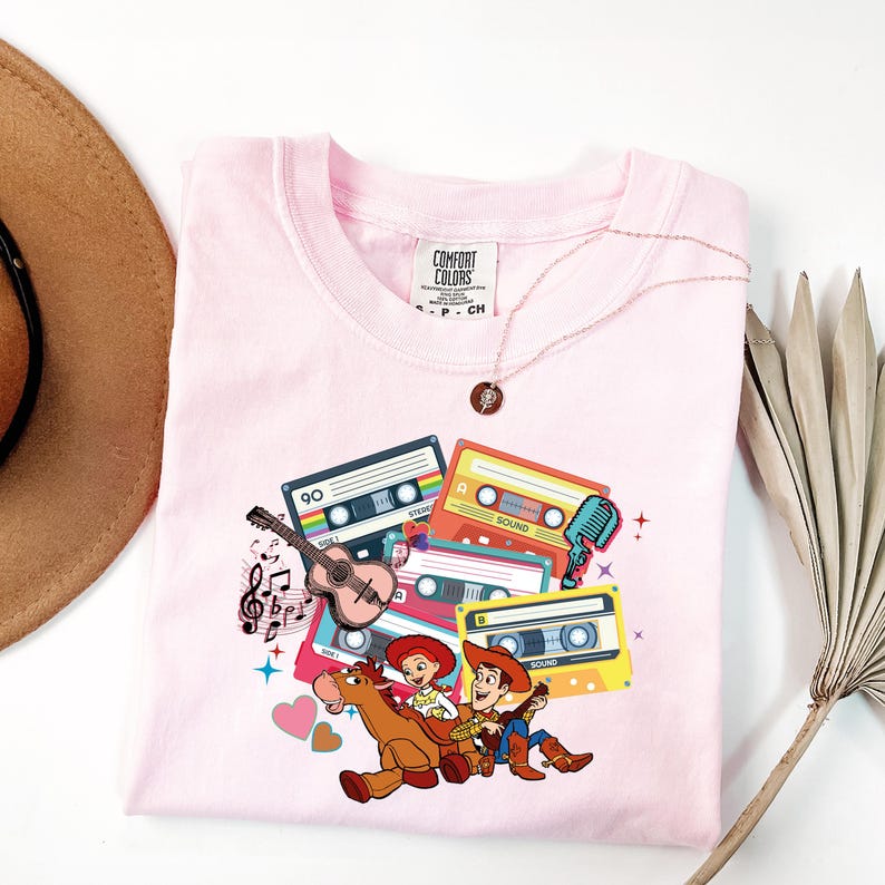 Puede incluir: Camiseta rosa Comfort Colors con un gr&aacute;fico de cintas de cassette, una guitarra, notas musicales y personajes de dibujos animados. Tambi&eacute;n se ven un collar dorado y un sombrero marr&oacute;n.