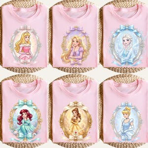 Op de afbeelding: Roze T-shirts met portretten van geanimeerde prinsessen in sierlijke frames. Elk shirt toont een andere prinses, waaronder Aurora, Rapunzel, Elsa, Ariel, Belle en Assepoester. De portretten zijn omgeven door decoratieve strikken.