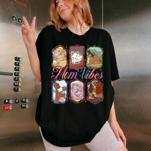 May include: Black t-shirt with the text "Mom Vibes" in a colorful script. The shirt features framed images of animated characters. The person in the image is wearing a black t-shirt and light-colored shorts.