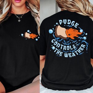 May include: Black t-shirt with a cartoon graphic of an orange fish with the text "Pudge Controls the Weather". The design is on the front and back of the shirt. The shirt has a crew neck and short sleeves.