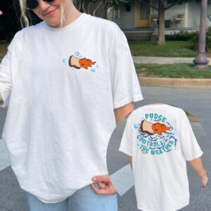 May include: White t-shirt with a cartoon design of an orange creature emerging from a brown object, surrounded by blue bubbles. The back of the shirt features the text "Pudge Controls The Weather" in blue.